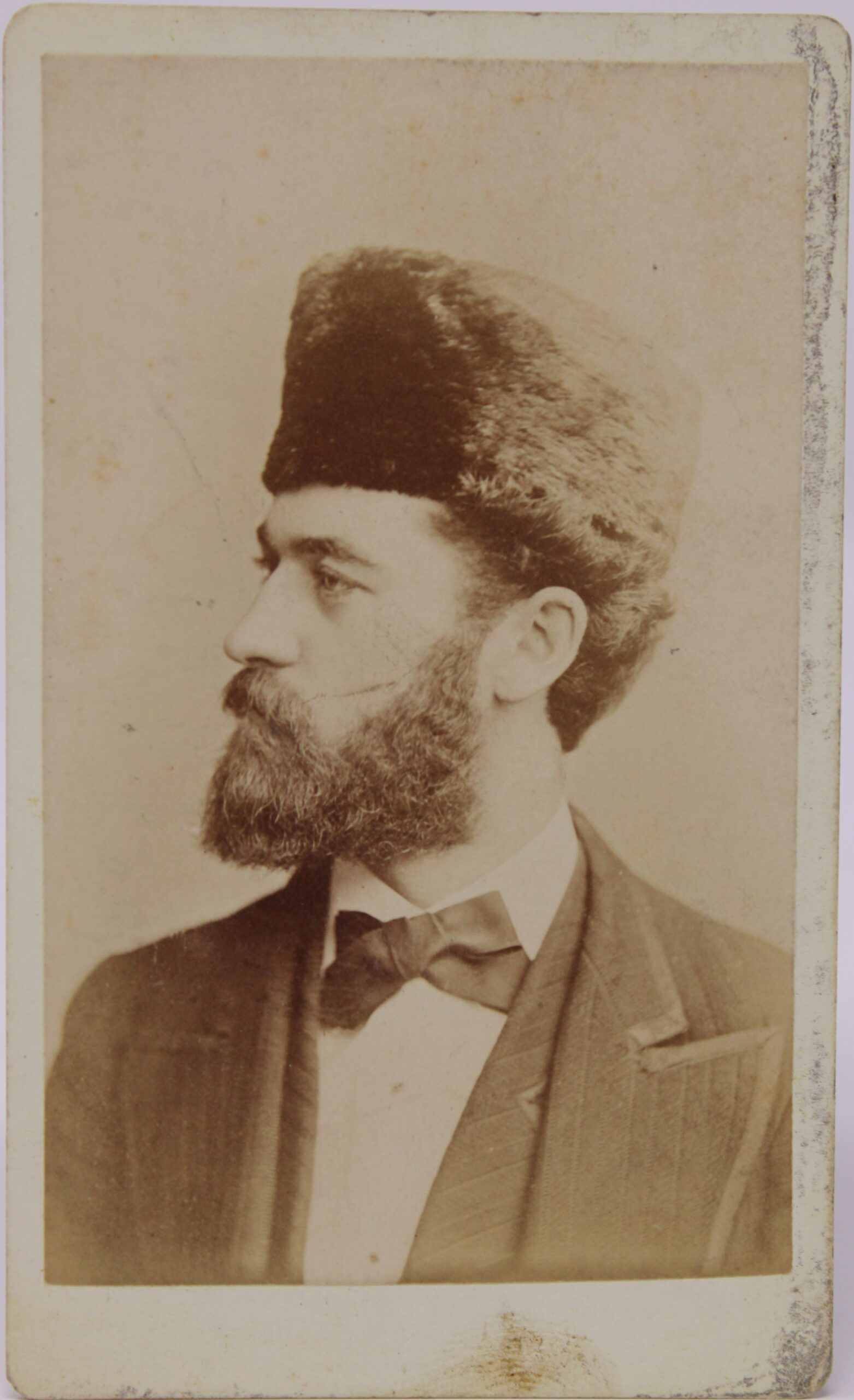 Vintage photogtaph in sepia showing side view of man with bow tie, full beard and fur hat.